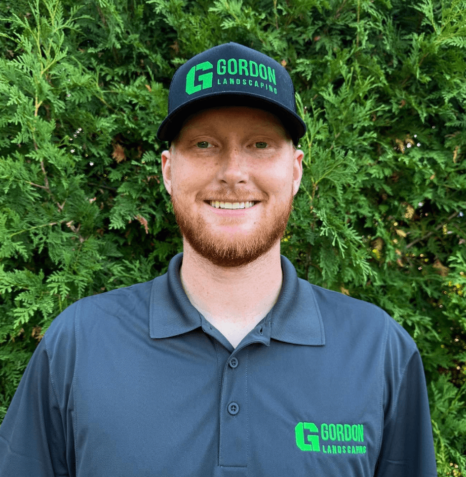 Smiling man with red beard wearing Gordon Landscaping apparel, posed against a green cedar hedge.